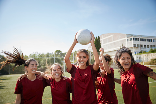 Female soccer player looking at camera celebrating goal with teammates in soccer field holding ball up