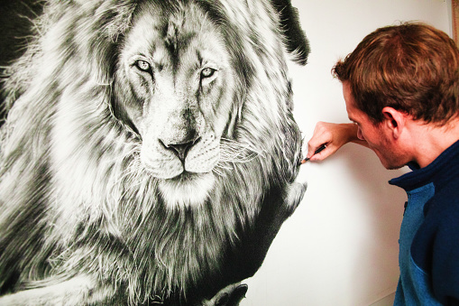 Over the shoulder view of a male artist drawing a lion with a charcoal pencil. The paper canvas is hanging on a wall.