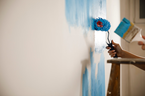 Copy space shot of unrecognizable couple using paintbrush and paint roller to apply blue paint on a white wall while renovating.