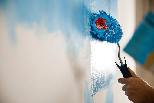 Close up shot of unrecognizable young woman applying blue paint on a white wall with a paint roller while renovating.