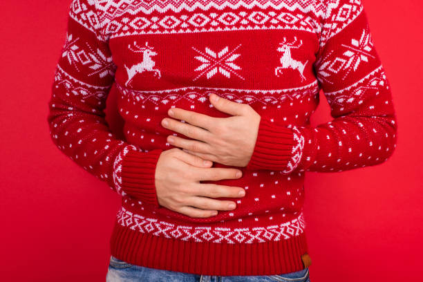 concepto de vientre. foto de cerca de los hombres con suéter rojo de invierno con ciervos sosteniendo su estómago aislado en el fondo rojo - christmas eating nausea illness fotografías e imágenes de stock