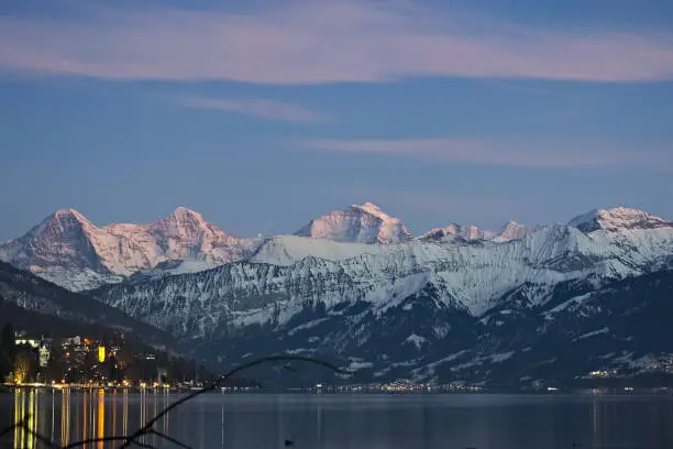 Famous mountain range Eiger, Moench and Jungfrau with snow on peaks at sunset reflecting in lake Thun..