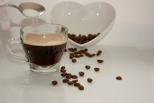 Spoon adding sugar substitute beans to cup with coffee and caffeine that in the background features a white heart-shaped bowl with coffee beans in Mexico City