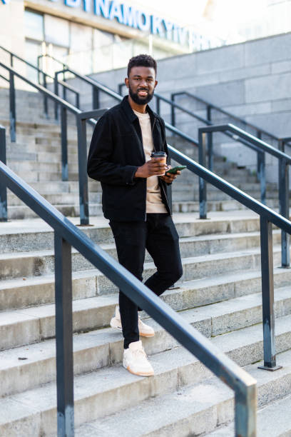 istock Handsome Afro American man is using a smart phone, drinking coffee and smiling while go down on stairs during city walk 1287294864