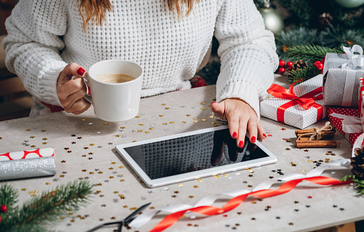 Unrecognizable woman holding coffee cup and using tablet at Christmas eve