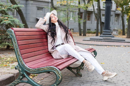 Beautiful woman in a coat and hat with pleasure sits on a bench on a city street