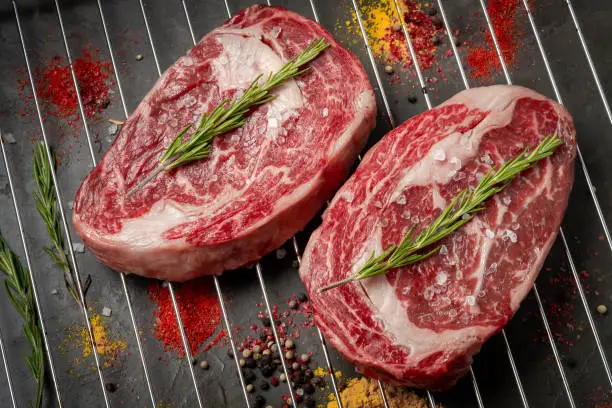 Two juicy raw rib eye steaks prepared for grilling with spices and herbs.