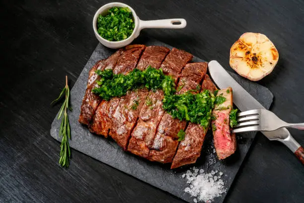 Ready to eat black Angus beef rib eye steak sliced with herbs, garlic and sauce on slate Board. Ready meal for dinner on the background of dark ebony.