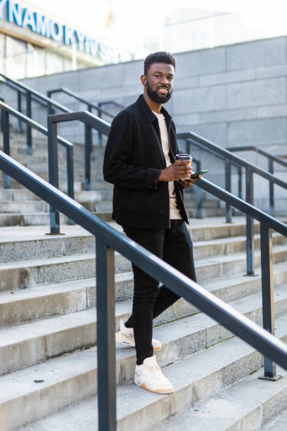 istock Handsome Afro American man is using a phone, holding a cup of coffee and smiling while walking in city 1287298820
