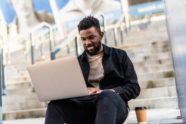 istock Young african cheerful man sitting on staircase and using laptop. 1287300286