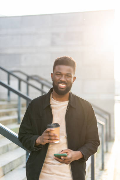 istock Handsome Afro American man is using a smart phone, drinking coffee and smiling while go down on stairs during city walk 1287303269