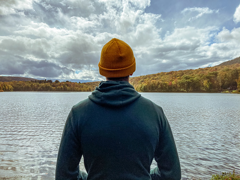 Young man only stands in contemplation looking at the view of a beautiful lake surrounded by trees with vibrant fall colors.