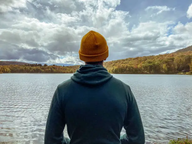 Photo of Young man only stands in contemplation looking at the view of a beautiful lake surrounded by trees with vibrant fall colors.