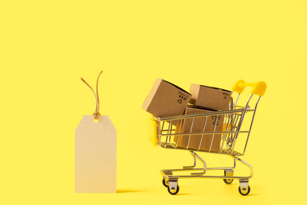 istock Toy shopping cart with boxes and empty tag on yellow background. Copy space for text or design. Sale, discount, shopping and delivery concept. Consumer society trend 1287312795