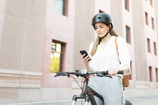 Good-looking woman texting on her smartphone outside an office building. Female worker using a helmet with a bicycle as an eco-friendly vehicle