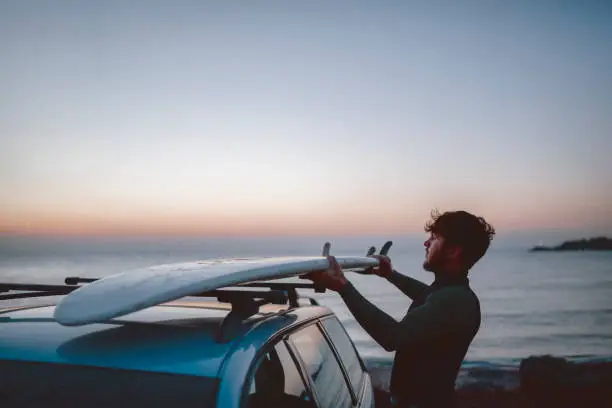 Side view of man unloading his surfboard from the car in the morning before sunrise.