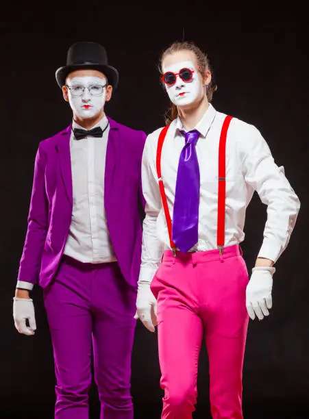 Portrait of male mime artists, isolated on black background. Two men wearing bright purple and pink suits go straight to the camera. Symbol of confident, brutal, cool.