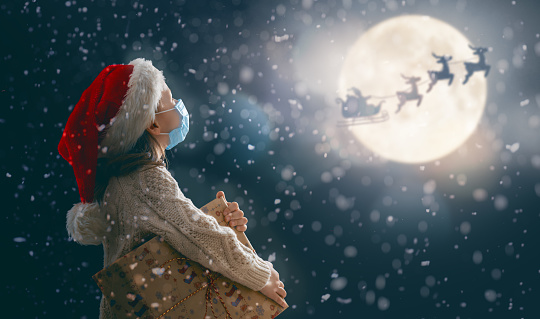 Merry Christmas! Cute little child with xmas present. Santa Claus flying in his sleigh against moon sky. Happy kid enjoy the holiday. Portrait of girl in face mask with gifts on dark background.