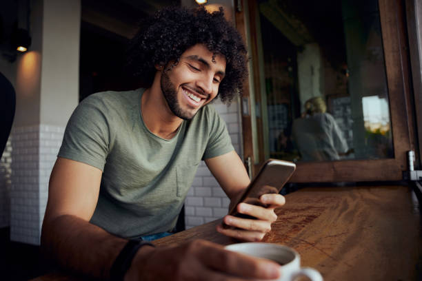 istock Handsome smiling young man sitting in cafe holding coffee mug while using smartphone 1287362582