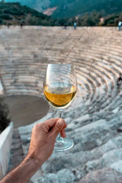 A glass of white wine in a man's hand. Greek Mediterranean wine against the backdrop of an antique amphitheater in Greece.