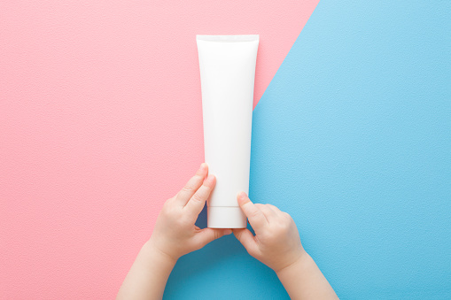 Baby little hands holding white cream tube on light pink blue table background. Pastel color. Care about clean and soft body skin. Daily children beauty product. Closeup. Point of view shot. Top view.