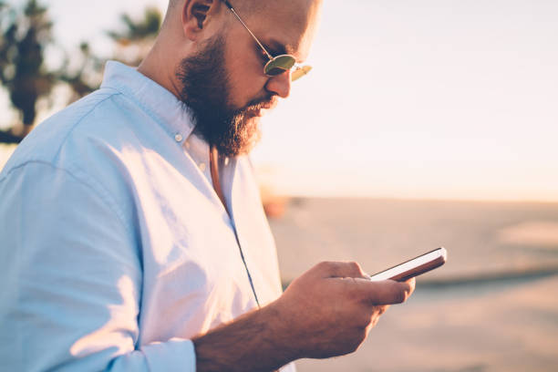 istock Bearded hipster guy holding mobile telephone making booking on website via application, close up of young man sending text message via smartphone chatting in social networks with friend on city street 1287373601