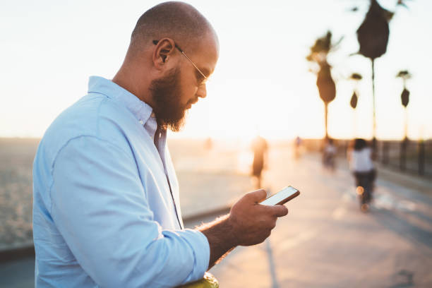 istock Young bearded male holding smartphone browsing information about cafes spending time on street, handsome hipster guy watching online video in social networks using mobile telephone during free time 1287373606