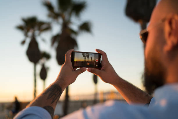 Back view of young man using smartphone camera for taking pictures of sunset and share in social networks, close up view of male blogger shooting video on cellphone of landscape during summer evening vista posterior del joven que utiliza la cámara del teléfono inteligente para tomar fotos de la puesta de sol y compartir en las redes sociales, vista de cerca de blogger masculino filmando video en el teléfono celular del paisaje durante la noche de ve