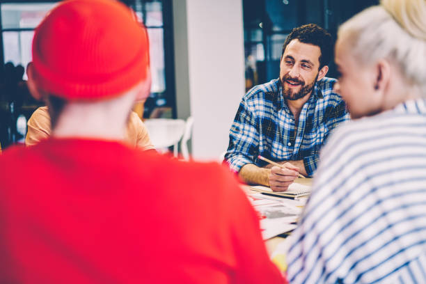 istock Positive young man dressed in smart casual wear laughing while discussing creative ideas and productive strategies with colleagues having brainstorming meeting sitting at table in modern office 1287374511