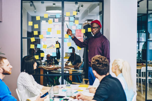 istock Smart casual dressed dark skinned young man conducting courses learning language for group of young people and pointing on colorful stickers with foreign words glued on glass wall in stylish coworking 1287374541