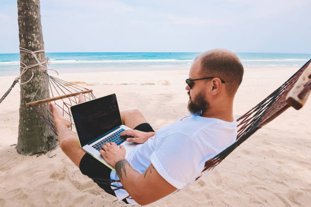 istock Male traveller in black sun glasses keyboarding on laptop computer doing freelance work while lying on hammock during summer vacation in tropical island. Mock up area on netbook, successful business 1287375010