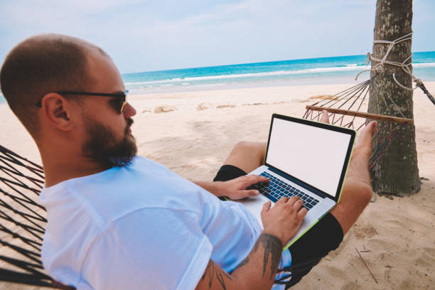 istock Pensive man freelancer in sunglasses texting on keyboard of modern laptop working remotely during summer vacations on sea shore. Male tourist businessman resting on hammock at distance job on computer 1287375015