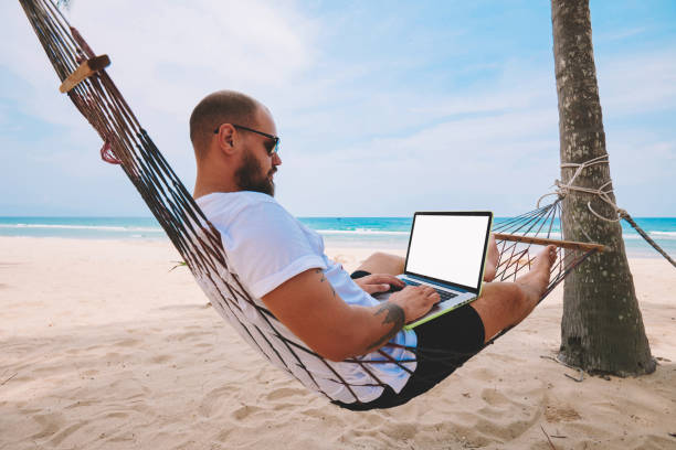 istock Young bearded businessman in sunglasses rest on hammock on ocean beach while working distantly on laptop computer with blank screen area for your web page advertise content during summer vacations 1287375016