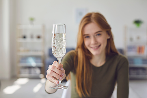 A young woman is drinking champagne. Focus on a glass of wine. A glass of champagne in a girl's hand in defocus indoors. Girl holds a glass of fart webcam video call. Date online.