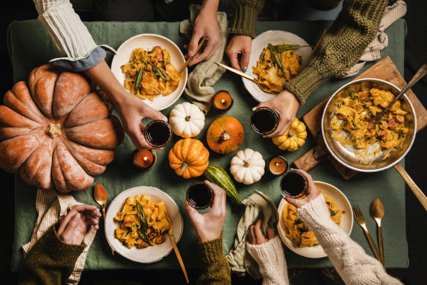 Friends celebrating Thanksgiving day with wine and squash pasta Flat-lay of Autumn dinner for gathering or Thanksgiving Day celebration. Friends clinking glasses with red wine and eating butternut squash pasta with sausage and sage over linen tablecloth, top view people banque stock pictures, royalty-free photos & images