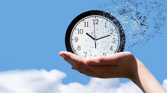 End of time or flight of time concept. Female hand holding a classic round clock falling into small pieces against a blue sky, front view, copy space