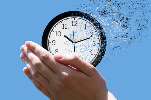 End of time or flight of time concept. Female hand closes the classic round clock falling into small pieces against the blue sky, front view, copy space