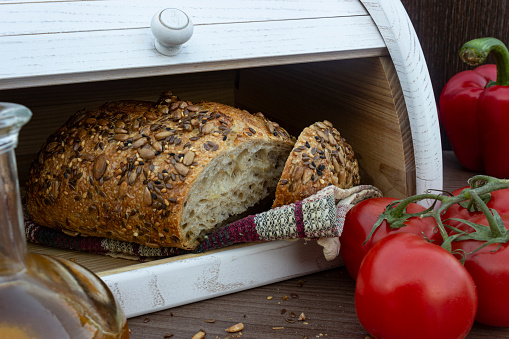 Fresh baked bread with seeds (sesame, sunflower seeds, linen) in white wooden bread box. Front view on tasty bread with olive oil, red vegetables and bread box on background.