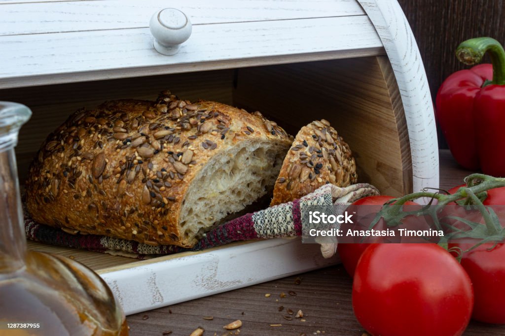 Close-up white wooden bread box with homemade bread inside. Breadbox with cutted bread, tomatoes, olive oil in glass bottle and red bell peppers on the dark wooden background. Fresh baked bread with seeds (sesame, sunflower seeds, linen) in white wooden bread box. Front view on tasty bread with olive oil, red vegetables and bread box on background. Bread Box Stock Photo