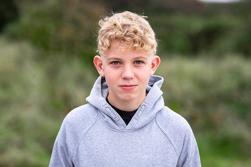 A portrait shot of a caucasian teenage boy in a rural outdoor setting looking at the camera with a contented emotion and wearing a hooded sweatshirt.