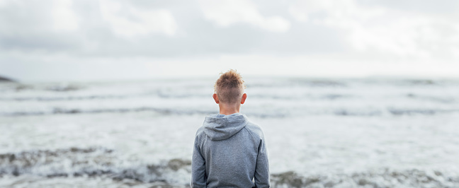 A rear view shot of an unrecognisable teenage boy wearing a hooded sweatshirt looking out to sea.