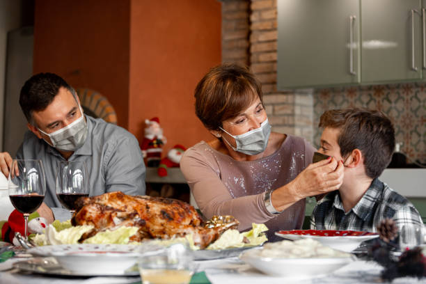 Grandmother adjust the surgical mask to her grandson during Christmas dinner. Family sitting at the Christmas Thanksgiving table at home in the year of the Coronavirus Pandemic. Grandmother adjust the surgical mask to her grandson. New rules, Covid outbreak, lockdown. thanksgiving holiday covid stock pictures, royalty-free photos & images