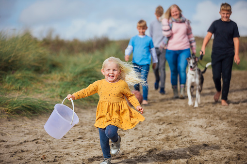 Young girl running along a beach holding a bucket in her hand on a sunny day in autumn. Her family are following along behind her with their family whippet dog. They are on a staycation to lower their carbon footprint and support the local economy.
