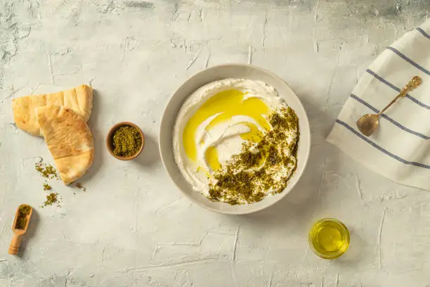 Photo of Popular middle eastern appetizer labneh or labaneh, soft white goat milk cheese with olive oil, hyssop or zaatar, served with pita bread on a grey table, top view,
