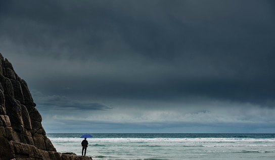 Man stood on rocks with umbrella at the coast, storm clouds overhead with clearer skies in the far distance.\nConcepts, weathering the storm.