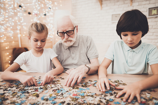 Grandfather, grandson and granddaughter at table at home. Grandpa and children are playing with puzzles.
