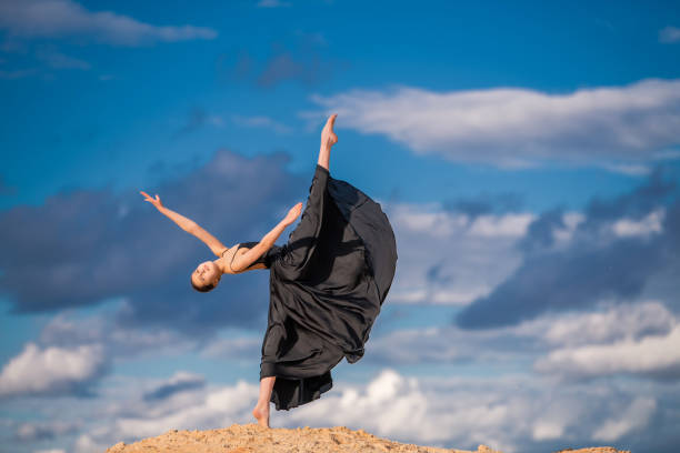 young ballerina in a black long dress hovers above the ground like a bird, resisting a gust of wind against a background of blue sky and clouds. The skirt develops like wings. young ballerina in a black long dress hovers above the ground like a bird, resisting a gust of wind against a background of blue sky and clouds. The skirt develops like wings. white crow stock pictures, royalty-free photos & images