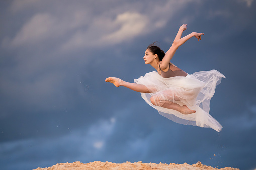 young ballerina in a light long white dress flies in a jump like a bird, a white skirt develops against the backdrop of a stormy evening sky. A rainbow appears among the clouds