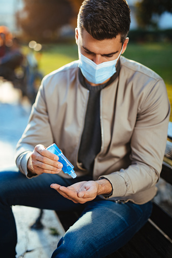 Mid adult man wearing protective face mask and disinfecting his hands with hand sanitizer outdoors