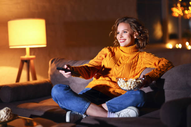 giovane donna allegra che mangia popcorn e guarda il film in tv mentre cambia canale con telecomando a casa solo la sera - home movie foto e immagini stock